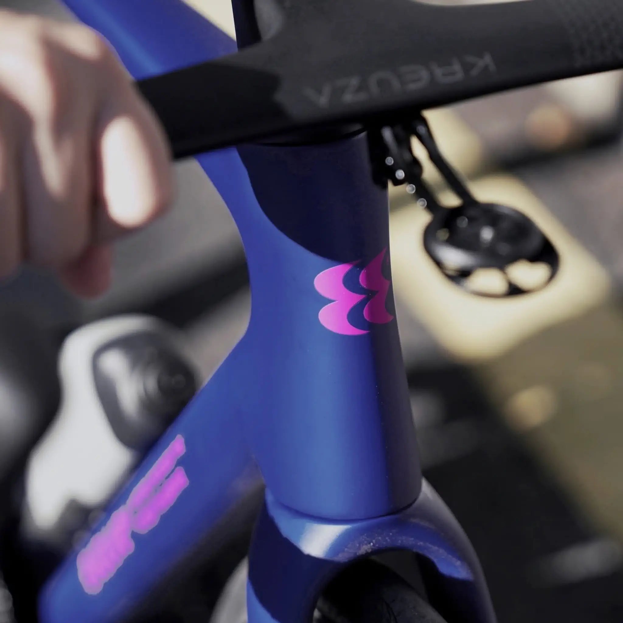 Close-up of a bicycle frame in a deep blue-purple color with a pink stylized logo, showing part of a rider's hand on the handlebar and a blurred outdoor background.