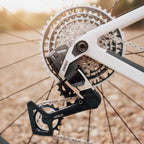 Close-up of a bicycle rear derailleur with a white frame and cassette in sharp focus against a blurred outdoor background.