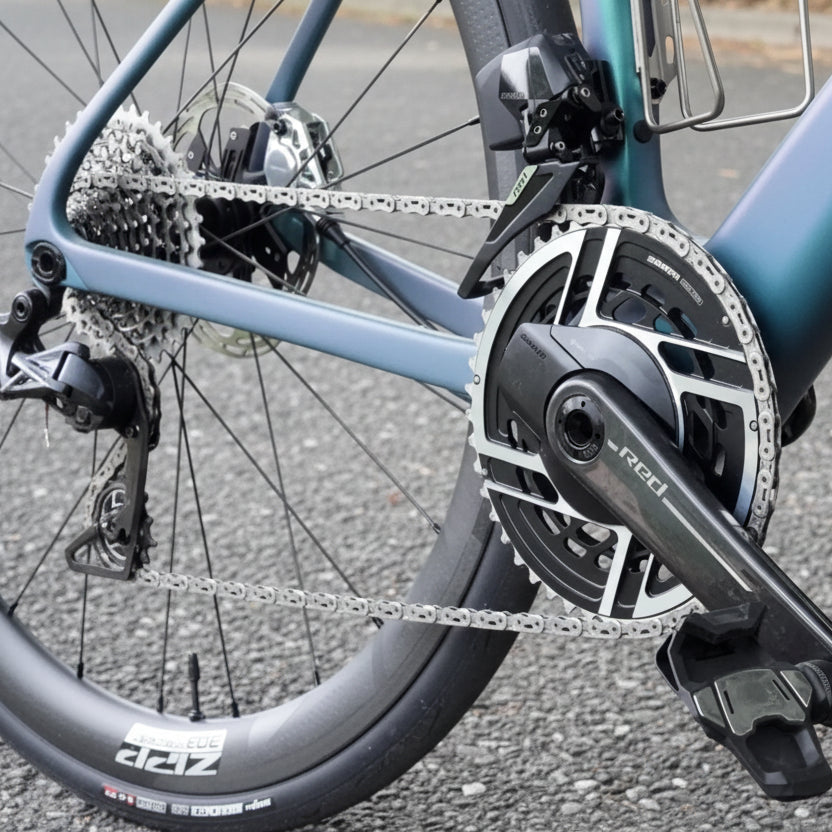 Close-up of a bicycle crankset with a black SRAM chainstay and silver chainrings, mounted on a teal bicycle frame outdoors among green grass and a tree trunk.